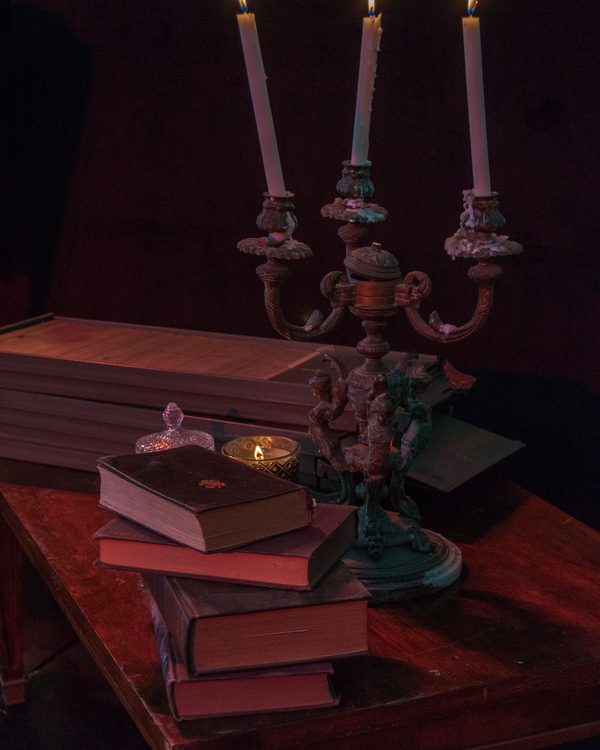 A tarnished candelabra with three lit candles on a red wooden table stacked with old books