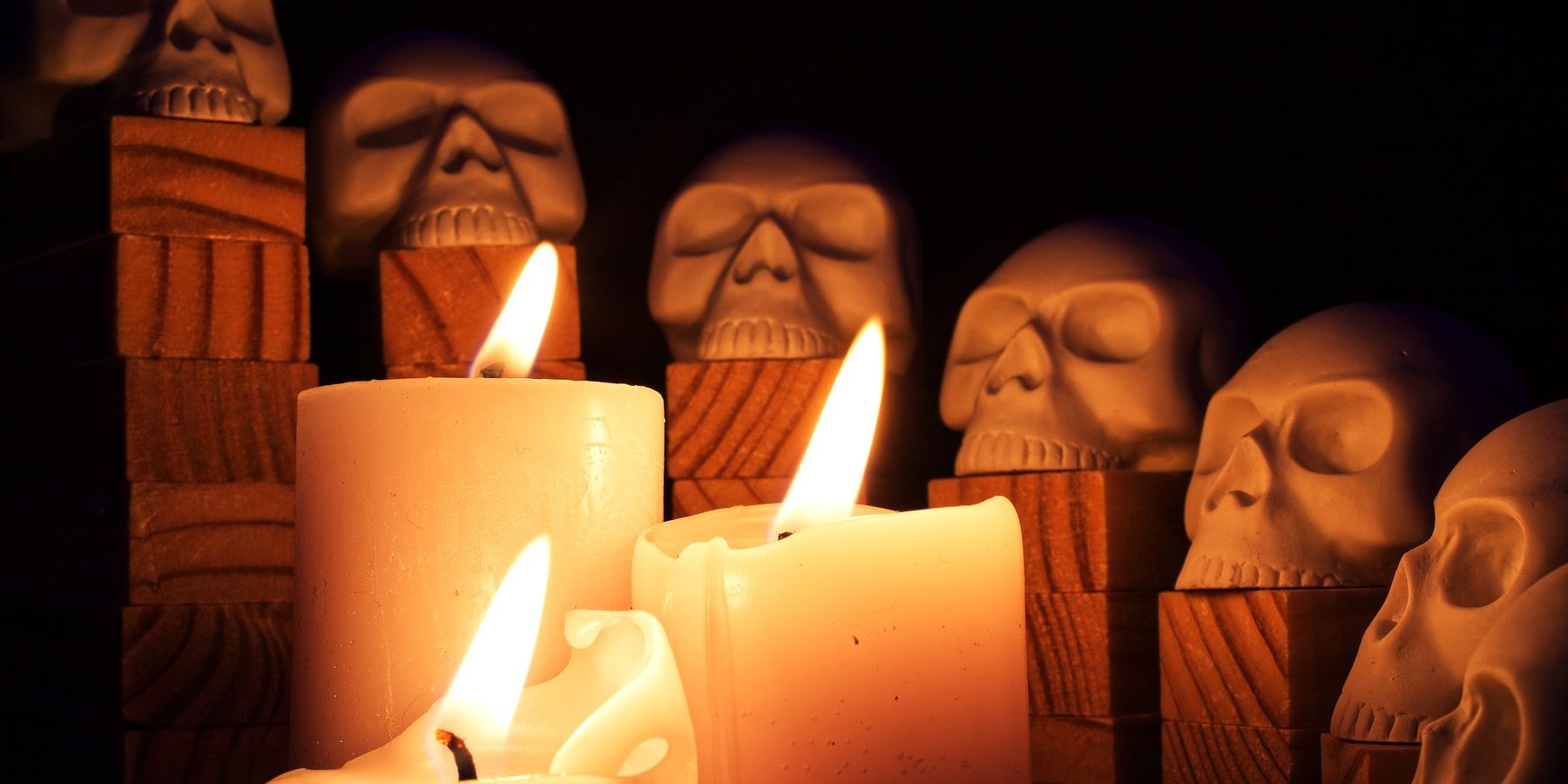 Pillar candles burning in front of a row of carved stone skulls on wooden blocks, the rest of the room in darkness