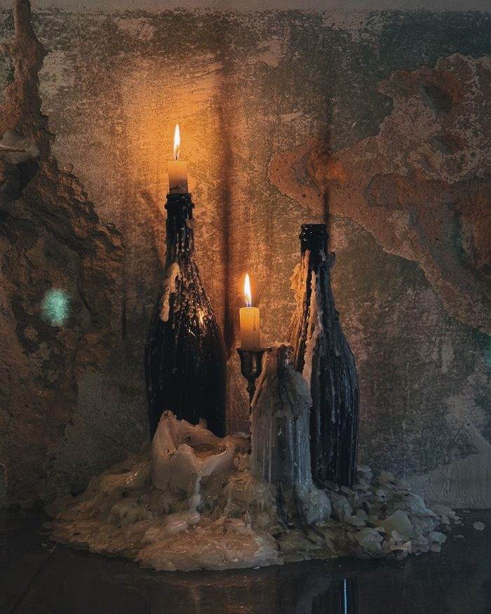 Two black wine bottles crusted in years of melted wax with candles burning in their necks, against a peeling plaster wall