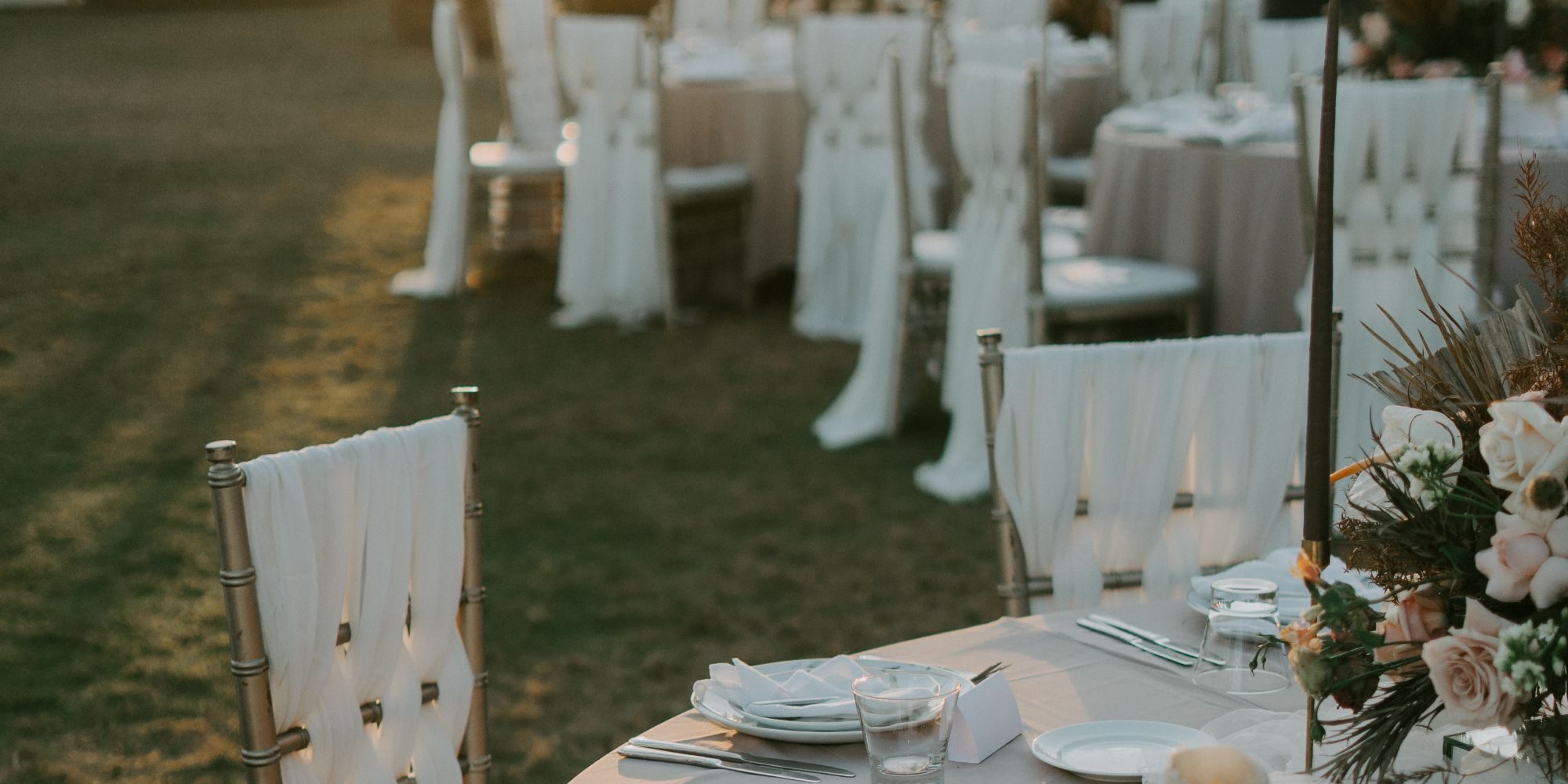 Long tables set for an outdoor dinner at golden hour, with linen, glassware and a low floral centerpiece