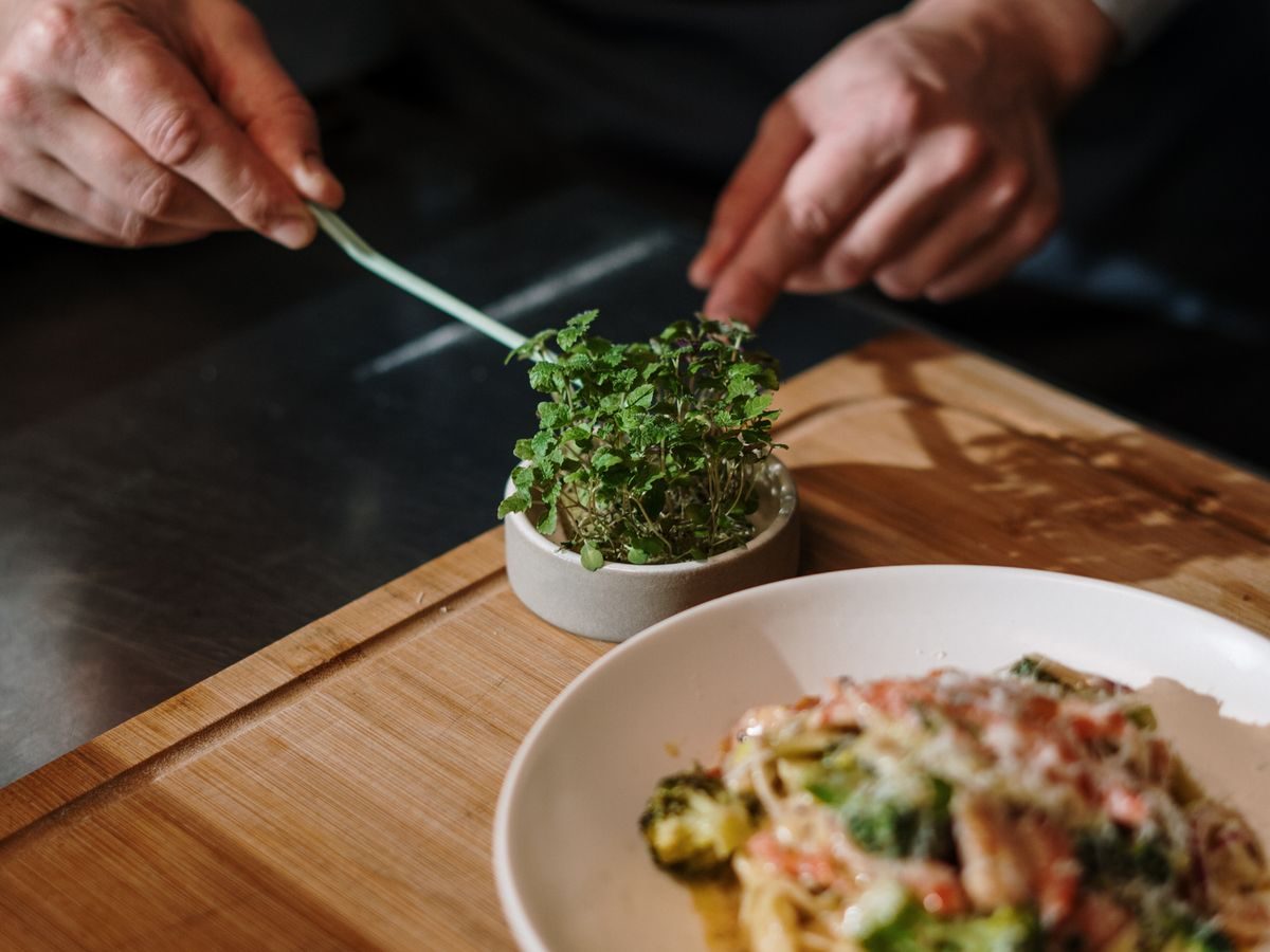 Hands finishing a bowl of pasta on a wooden board with fresh herbs alongside