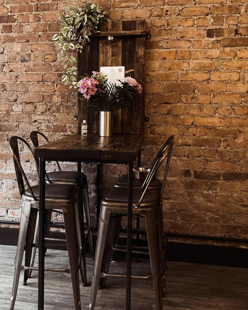 A dark wood high top table and four metal stools against an old brick wall, with a hanging plant and a vase of pink flowers