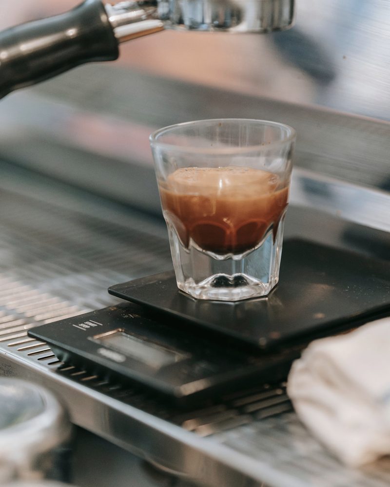 A shot of espresso in a small glass sitting on a black scale under the group head