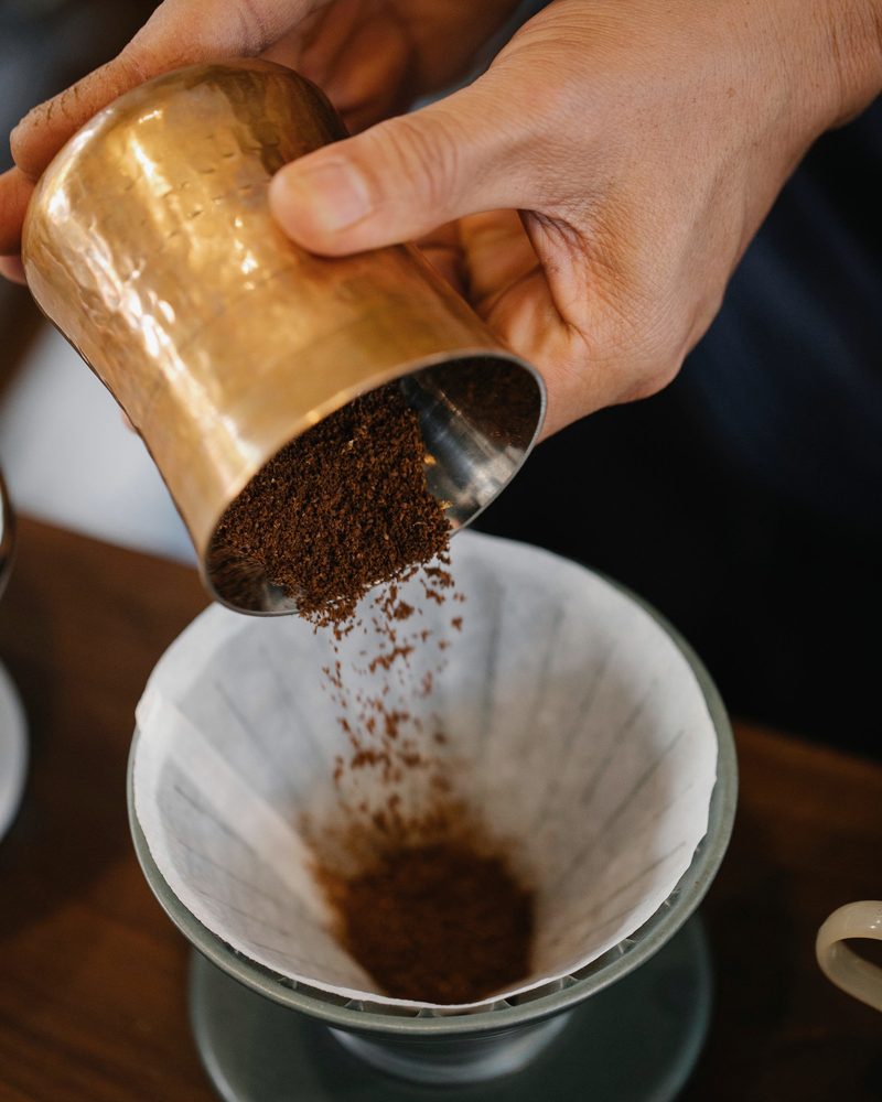 Hands tipping fresh grounds from a brass cup into a paper filter in a green ceramic dripper