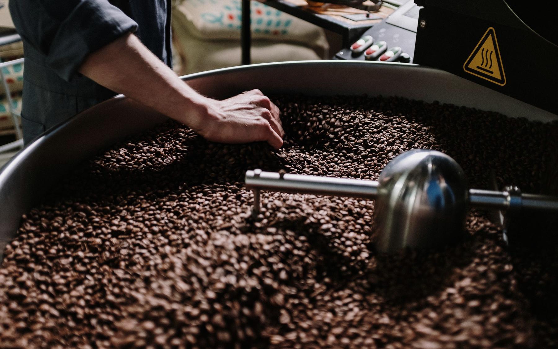 A hand turning fresh roasted coffee beans in the round cooling tray of a drum roaster, the steel stirring arm in the foreground