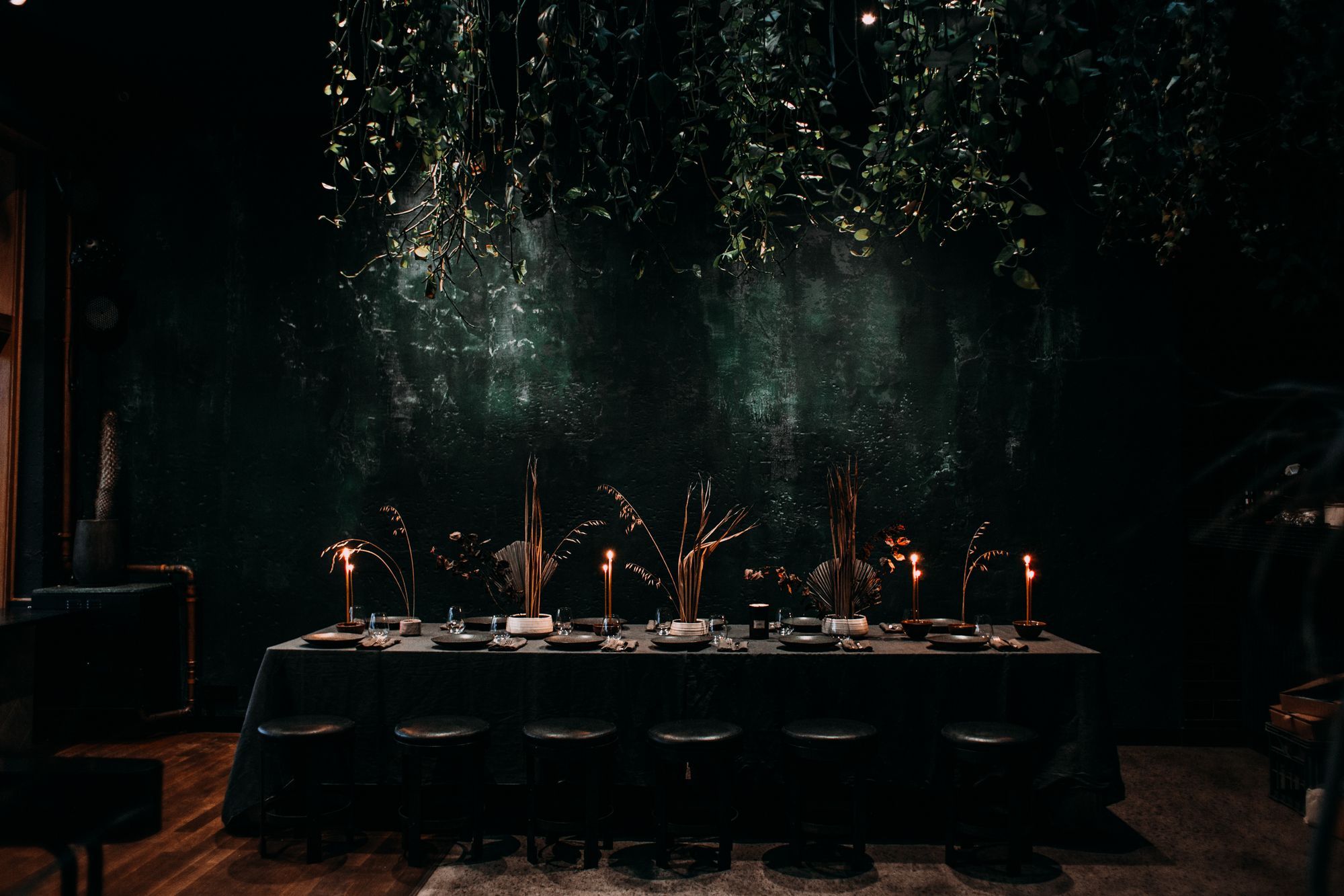 A long table set for dinner against a dark green plaster wall, lit by tall candles among dried grasses