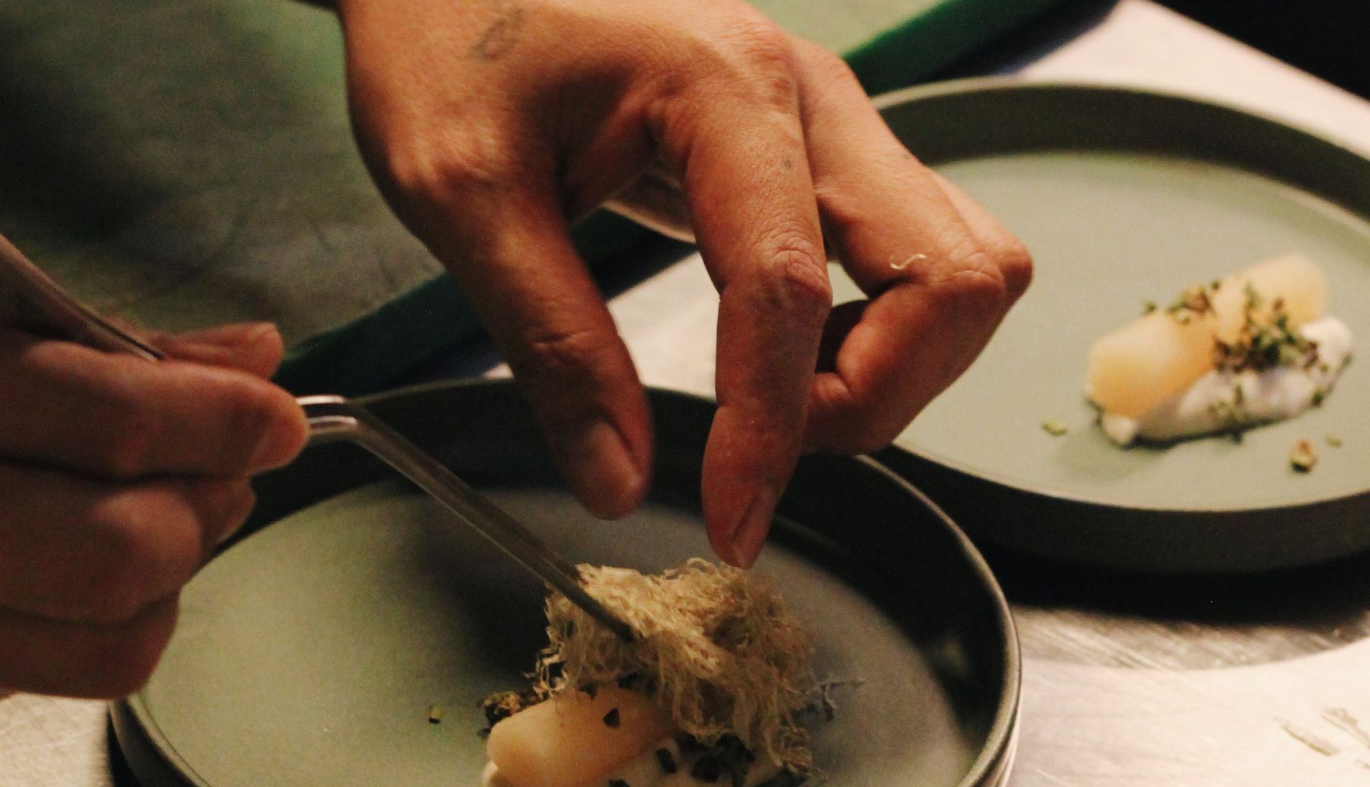 A cook placing a crisp garnish on a small green plate with tweezers at a steel kitchen counter