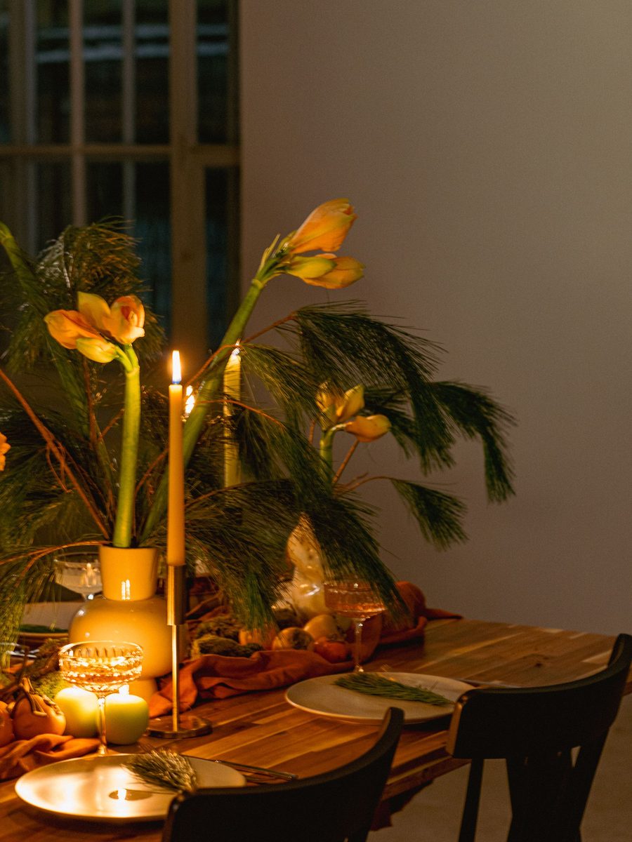 A candlelit wooden table with amaryllis and pine in a yellow vase, set with plates and cut glass coupes