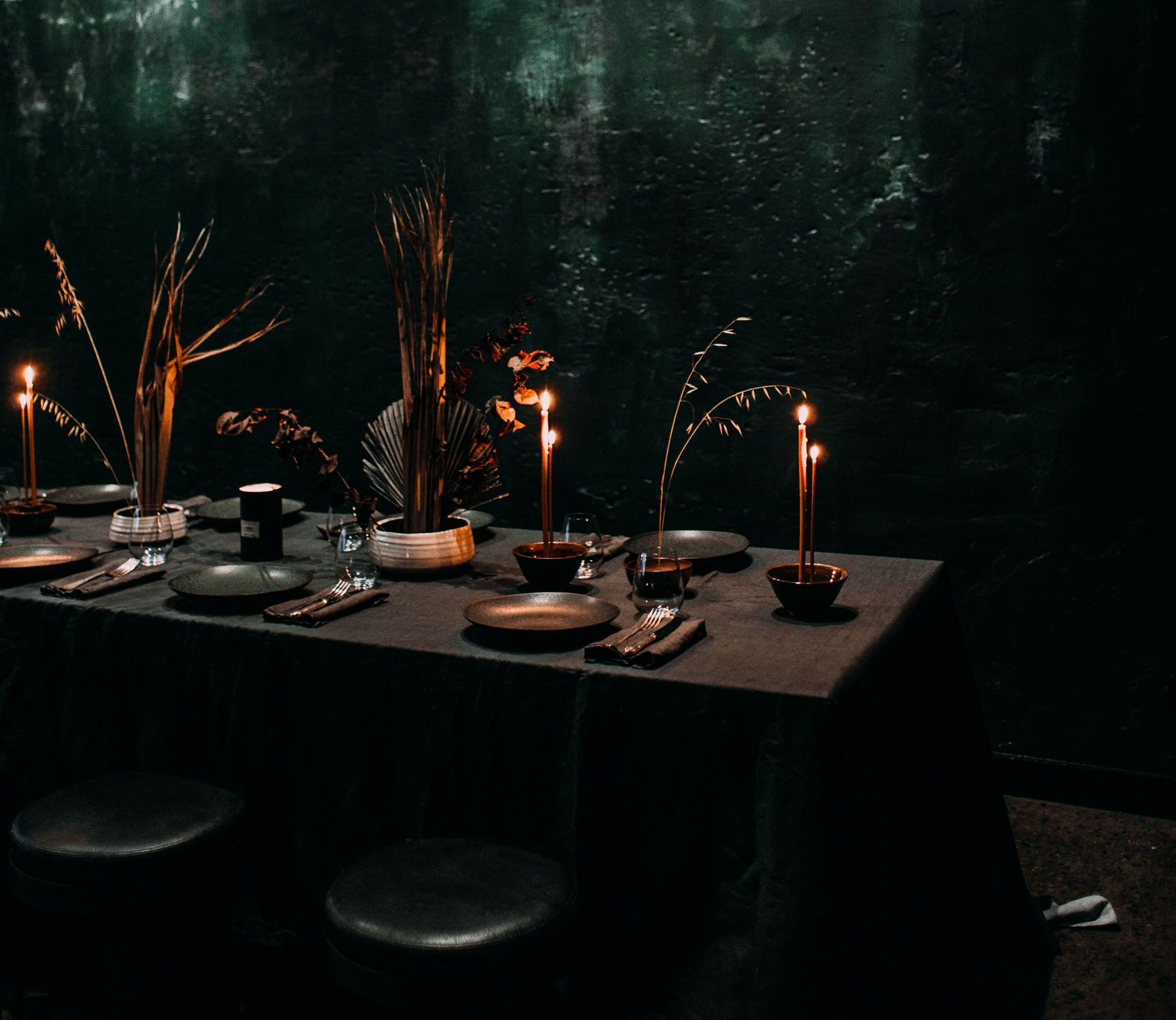A long table dressed in dark linen with tapered candles, dried grasses and hand thrown plates