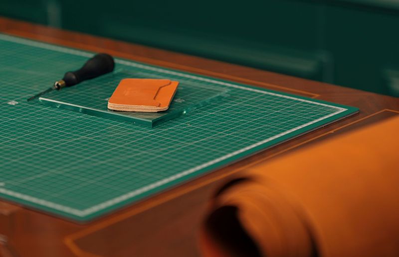 A tan card holder on a green cutting mat next to an awl and a roll of cognac leather