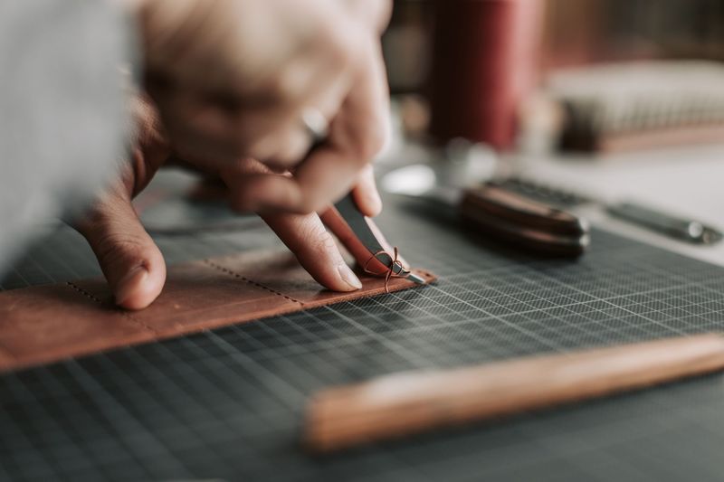 Two hands pulling waxed thread through a line of stitching holes in a strip of tan leather on a dark cutting mat