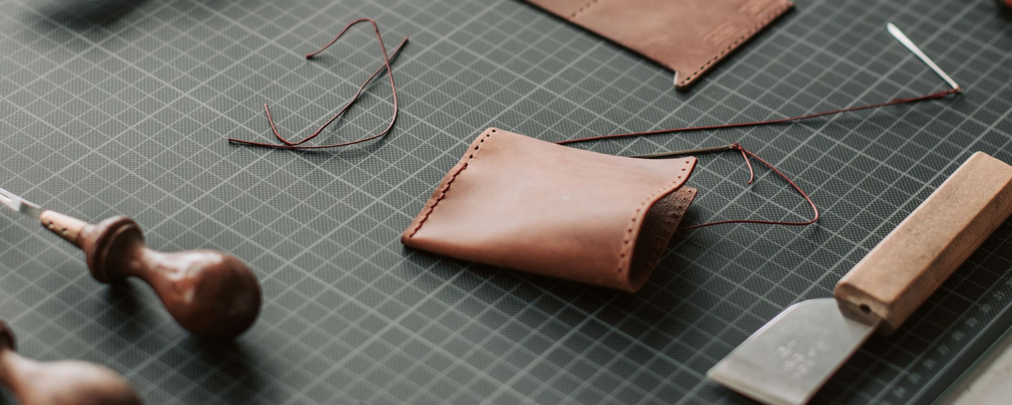A half stitched tan leather pouch on a grey cutting mat, threaded with red waxed linen, beside wooden handled tools and a skiving knife