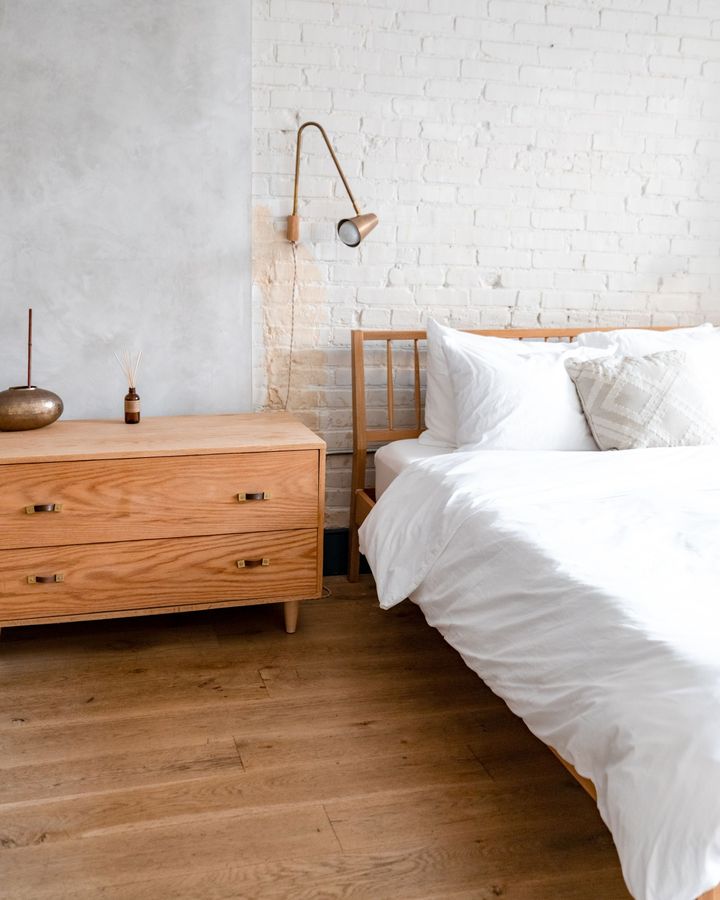 A freshly made bed in crisp white linen beside a wooden dresser and a white brick wall, ready for the next guest