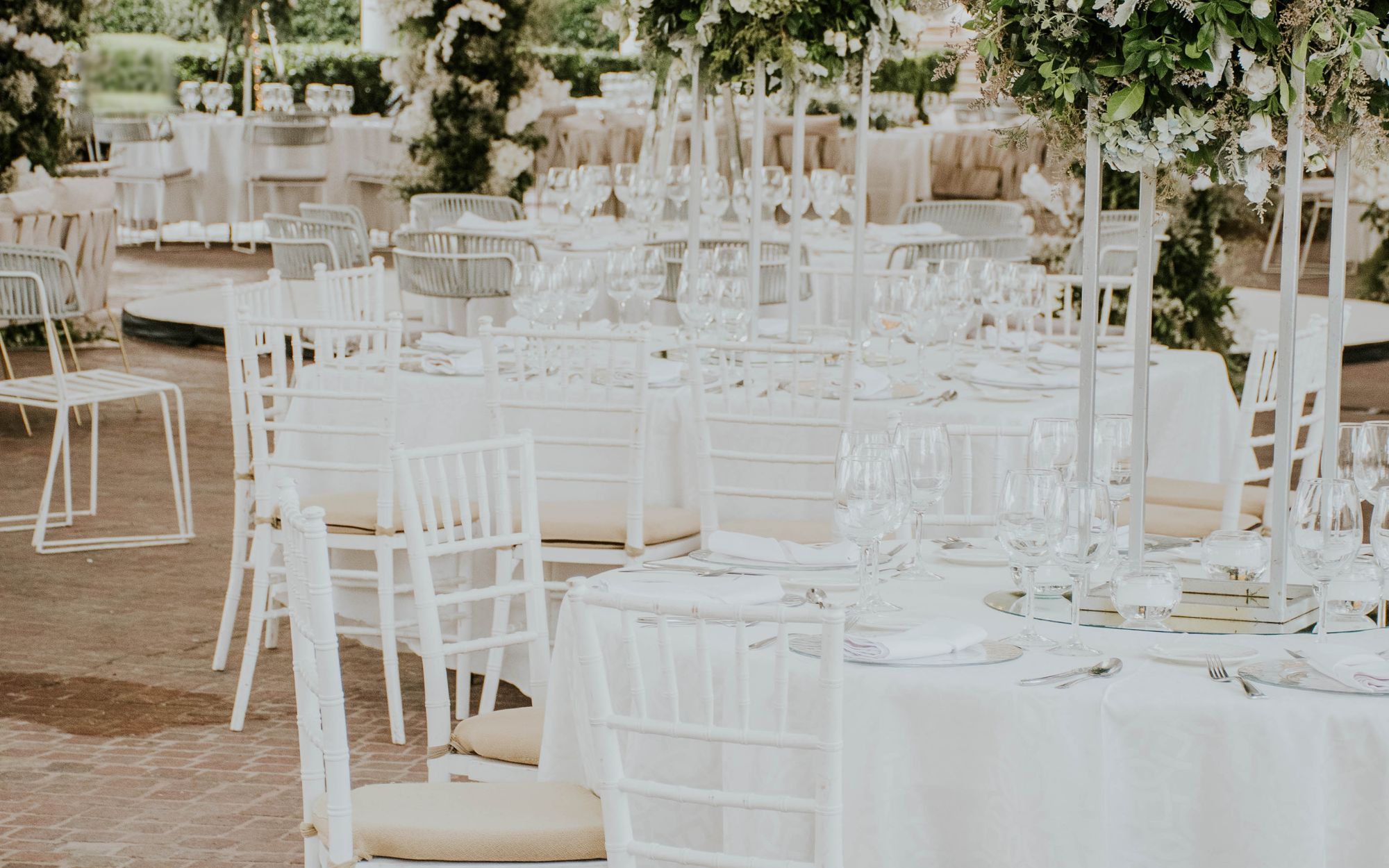 An elegant garden venue with white chairs tucked under round tables in crisp white linen beneath tall arrangements of white flowers and greenery