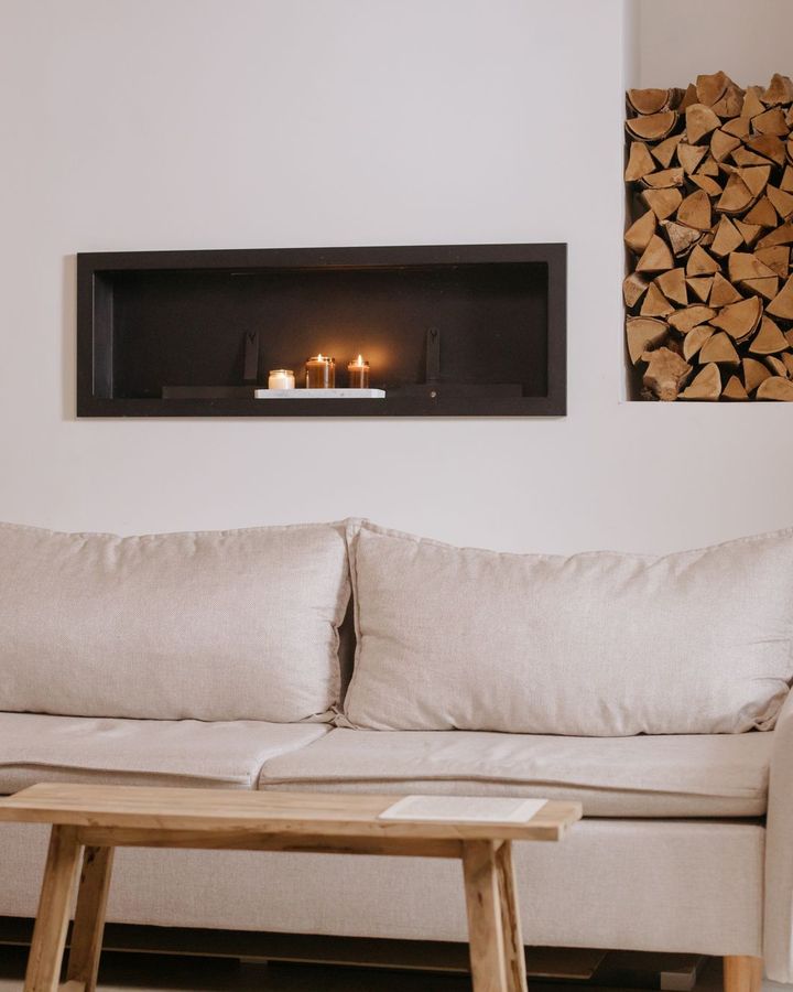 A cream linen sofa and a rustic wooden bench in a calm white living room with candles glowing in a wall niche