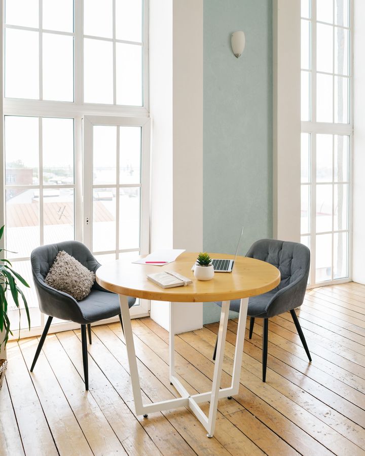 A sunlit office corner with tall windows, a round wooden table and two gray chairs on a clean plank floor