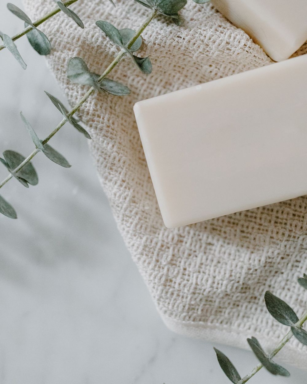 A plain white bar of soap resting on a woven cotton towel with sprigs of eucalyptus on pale marble