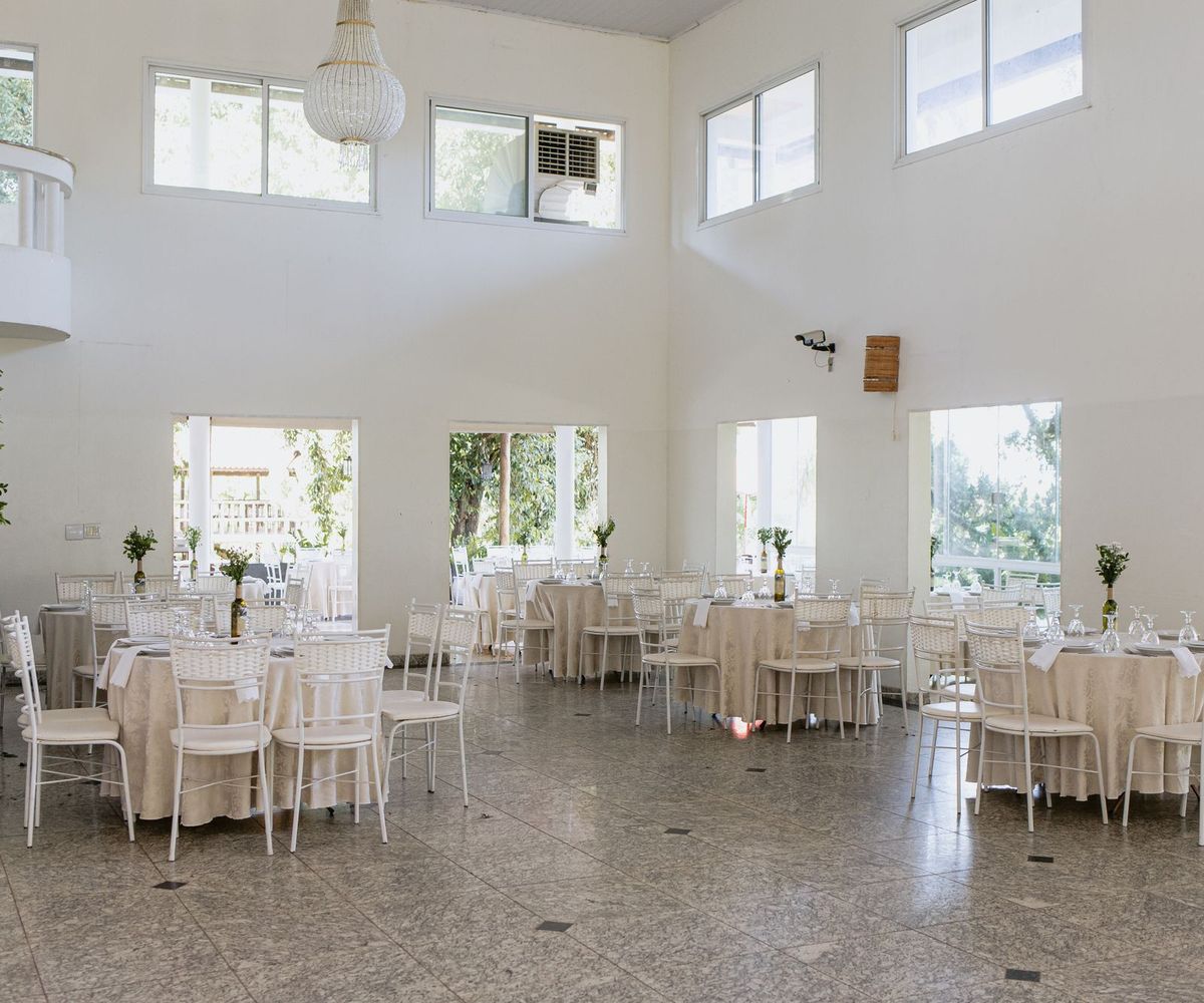 A bright, high ceilinged event hall with round tables in cream linen, white chairs and tall windows letting in daylight