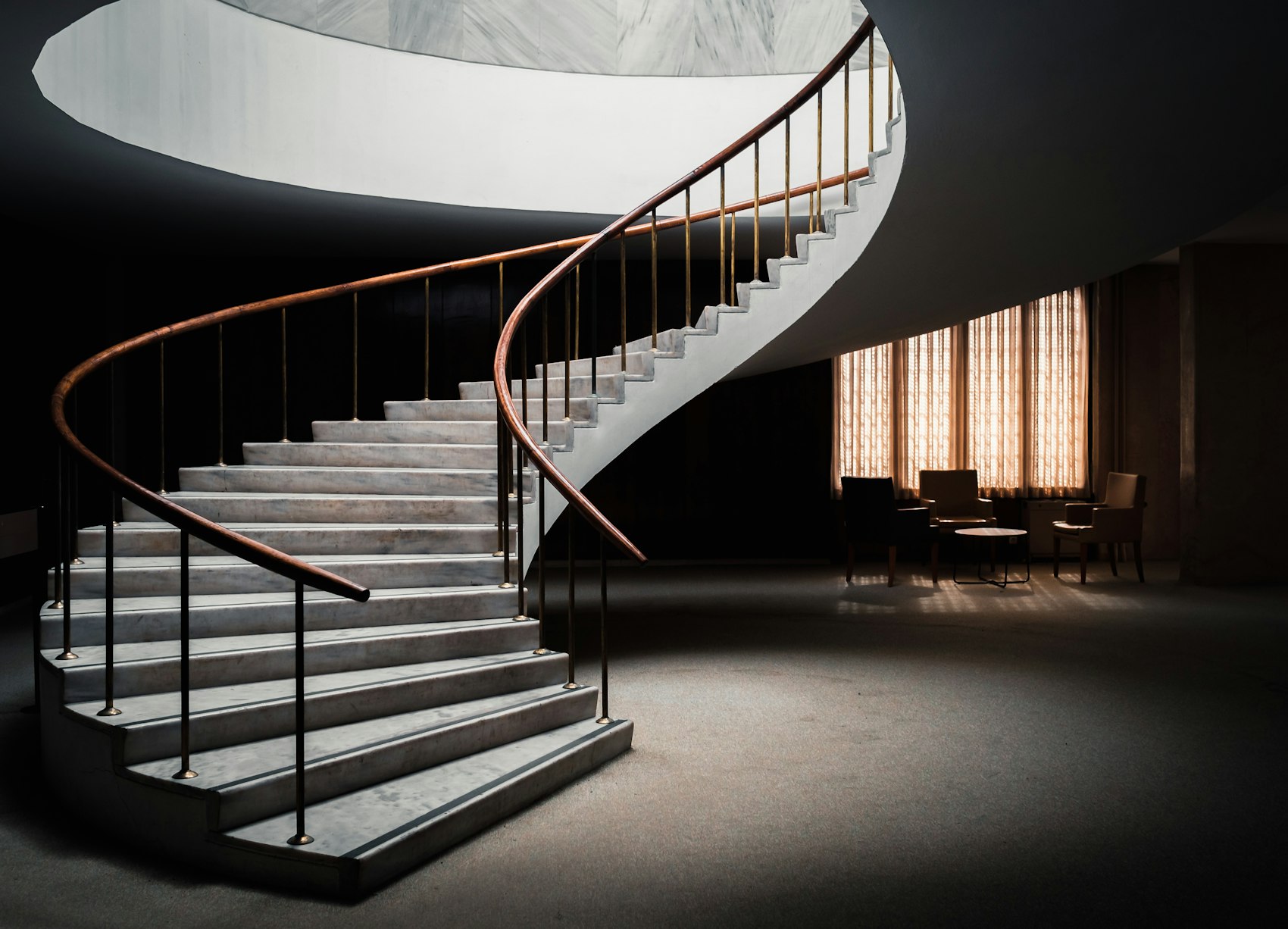 A pale spiral staircase curving down through a dark, double height entrance hall, lit by a low warm lamp.