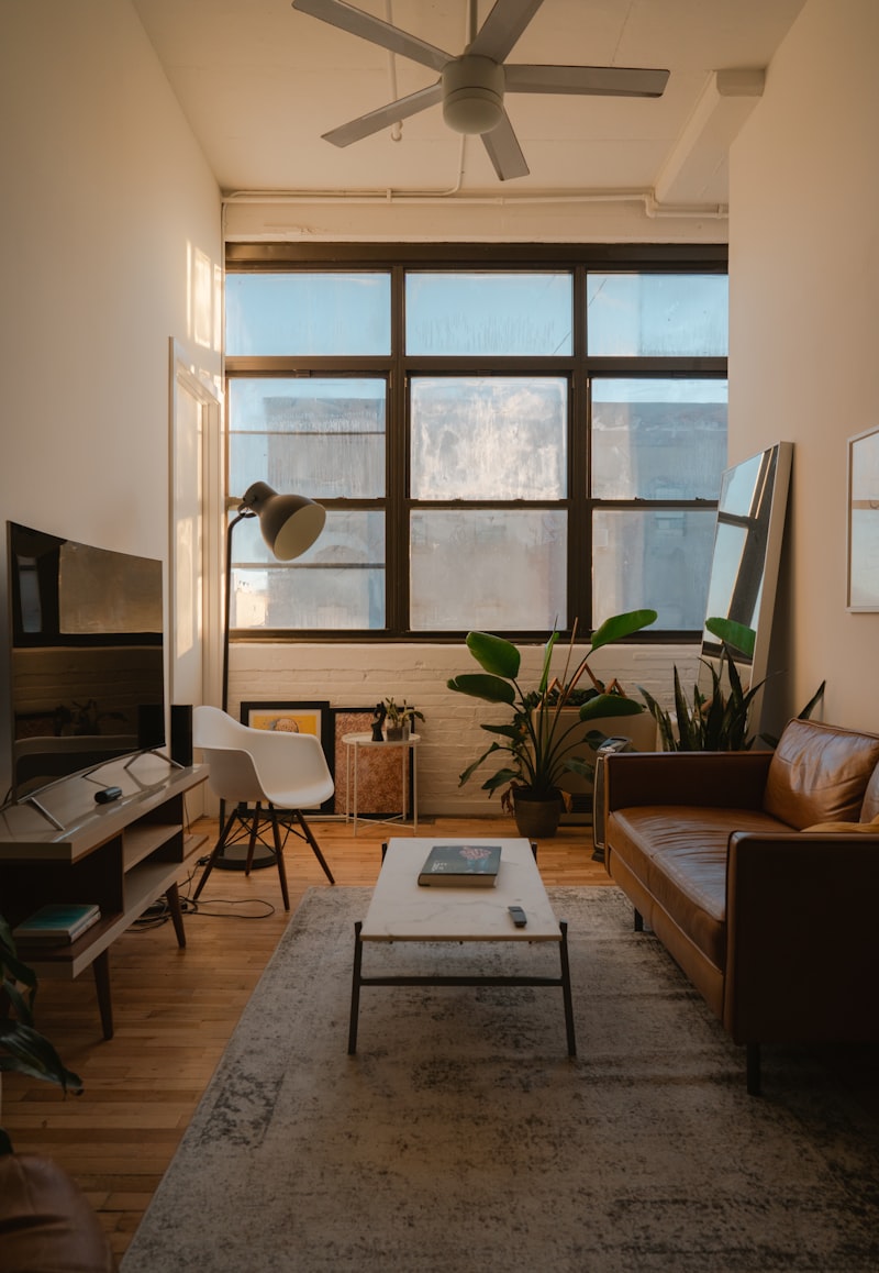 A warm loft interior with tall steel framed windows, a leather sofa, timber floors and a large plant.