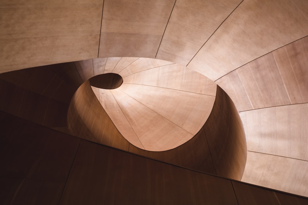 A warm timber spiral stair seen from directly above, its treads winding to a dark opening at the center.