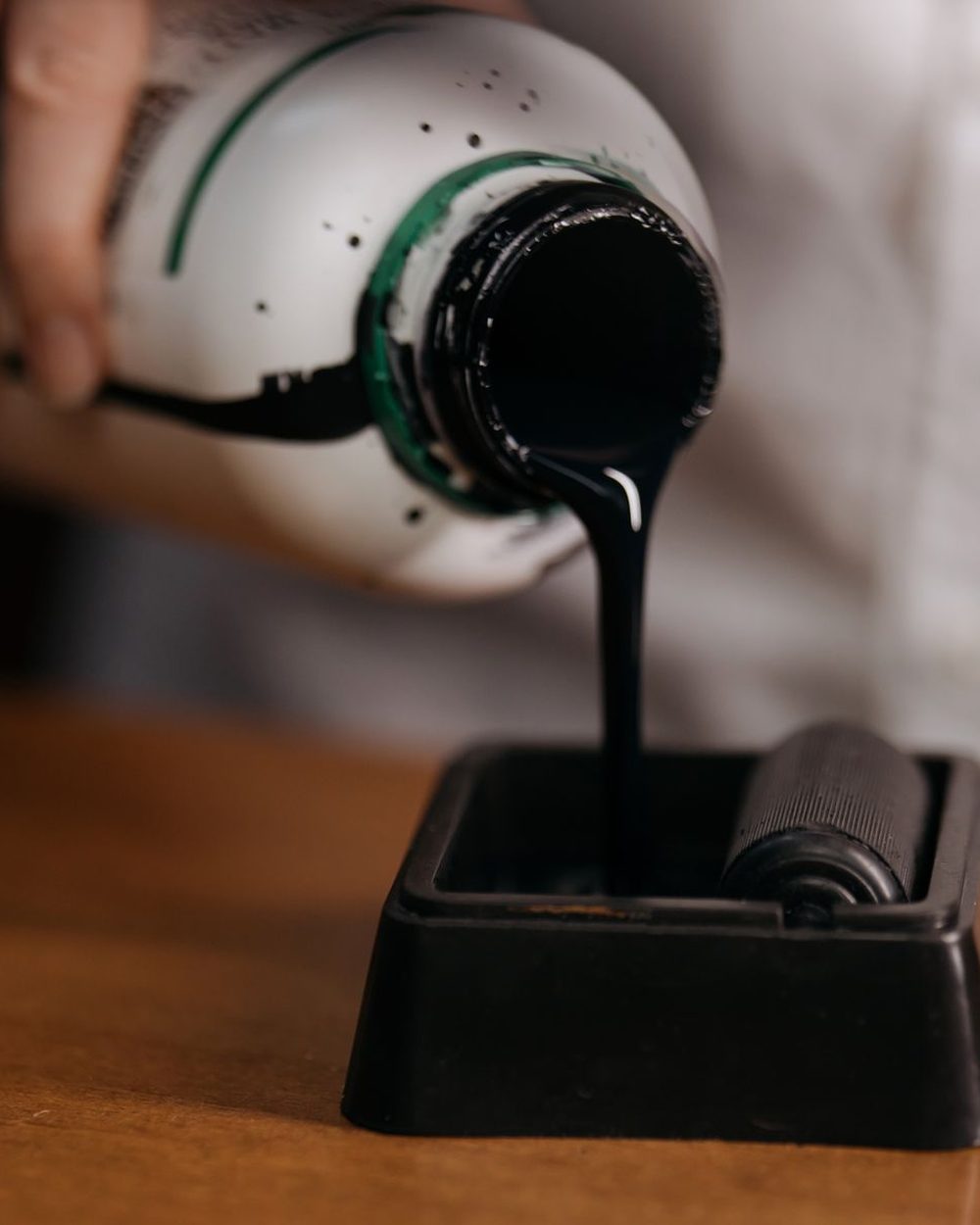 A hand pouring thick black ink from a white bottle into a small tray with a roller