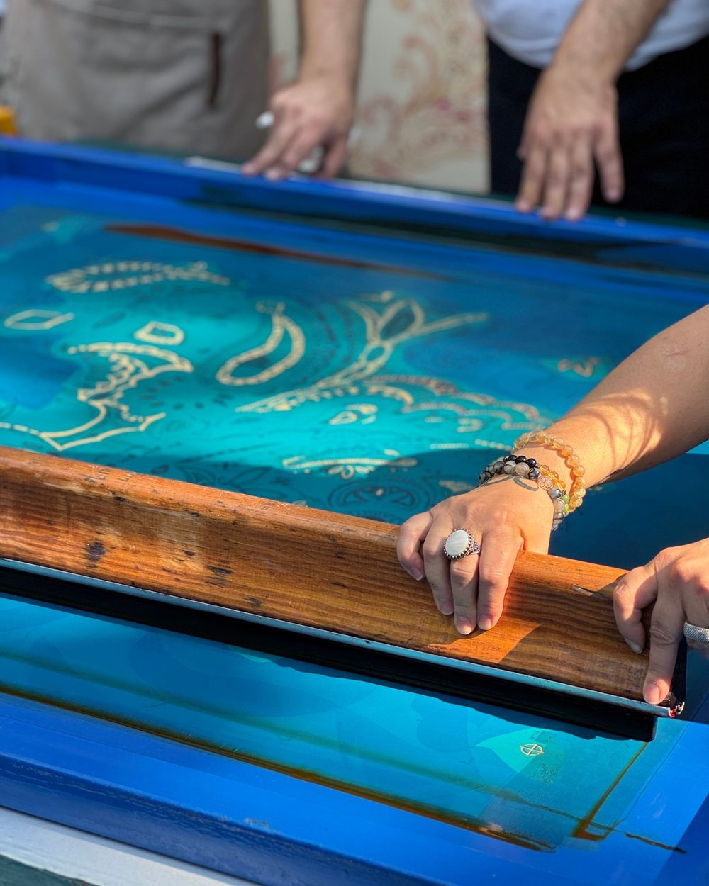 Two hands, one wearing a white stone ring and beaded bracelets, pulling a long wooden squeegee across a screen flooded with turquoise ink