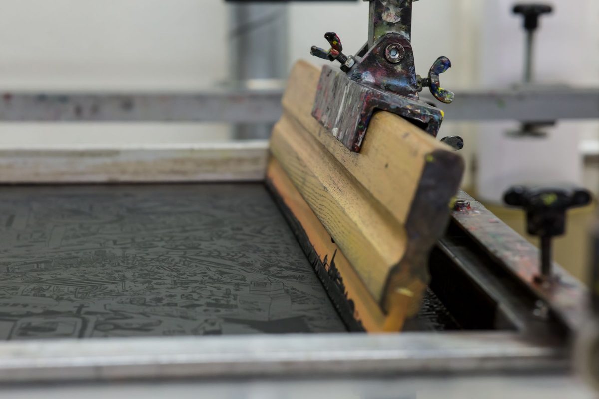 A wooden squeegee clamped into the arm of a screen printing press, resting on a used mesh screen