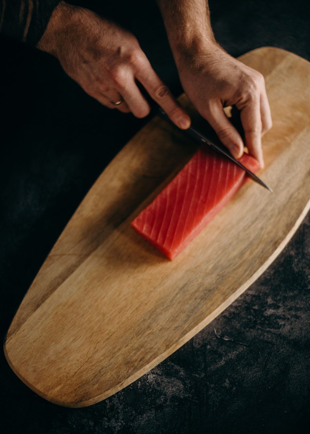 A chef's hands slicing a block of red tuna with a long knife on an oval wooden board