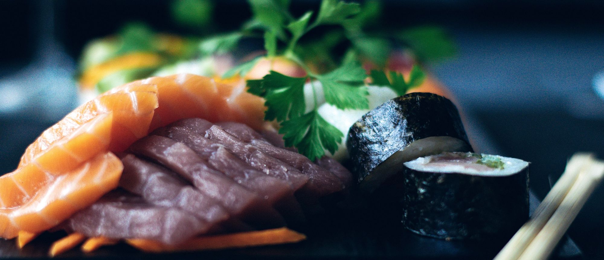 Folded slices of salmon and tuna sashimi with parsley and a cut maki roll on a black tray, chopsticks resting at the edge