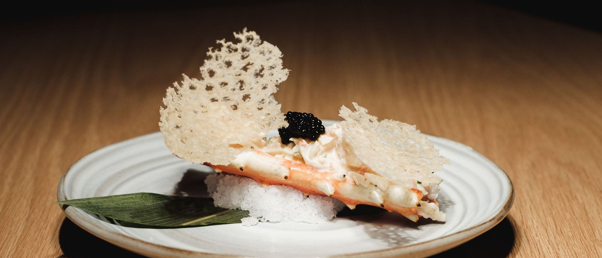A single omakase course of king crab and caviar with a lace rice cracker, on a small plate under a spotlight at a pale wood counter