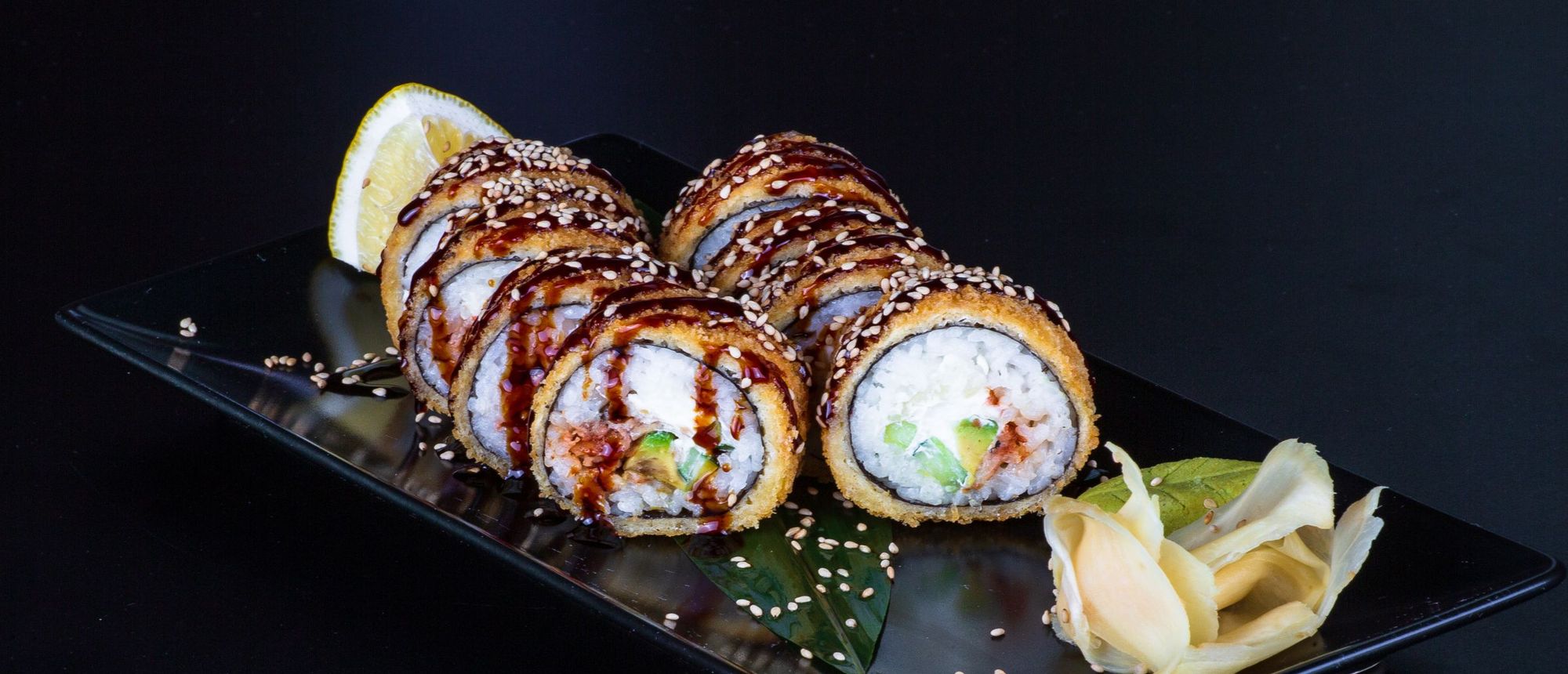 Sliced rolls topped with sesame and a drizzle of sweet soy glaze, with pickled ginger and lemon, on a long black plate