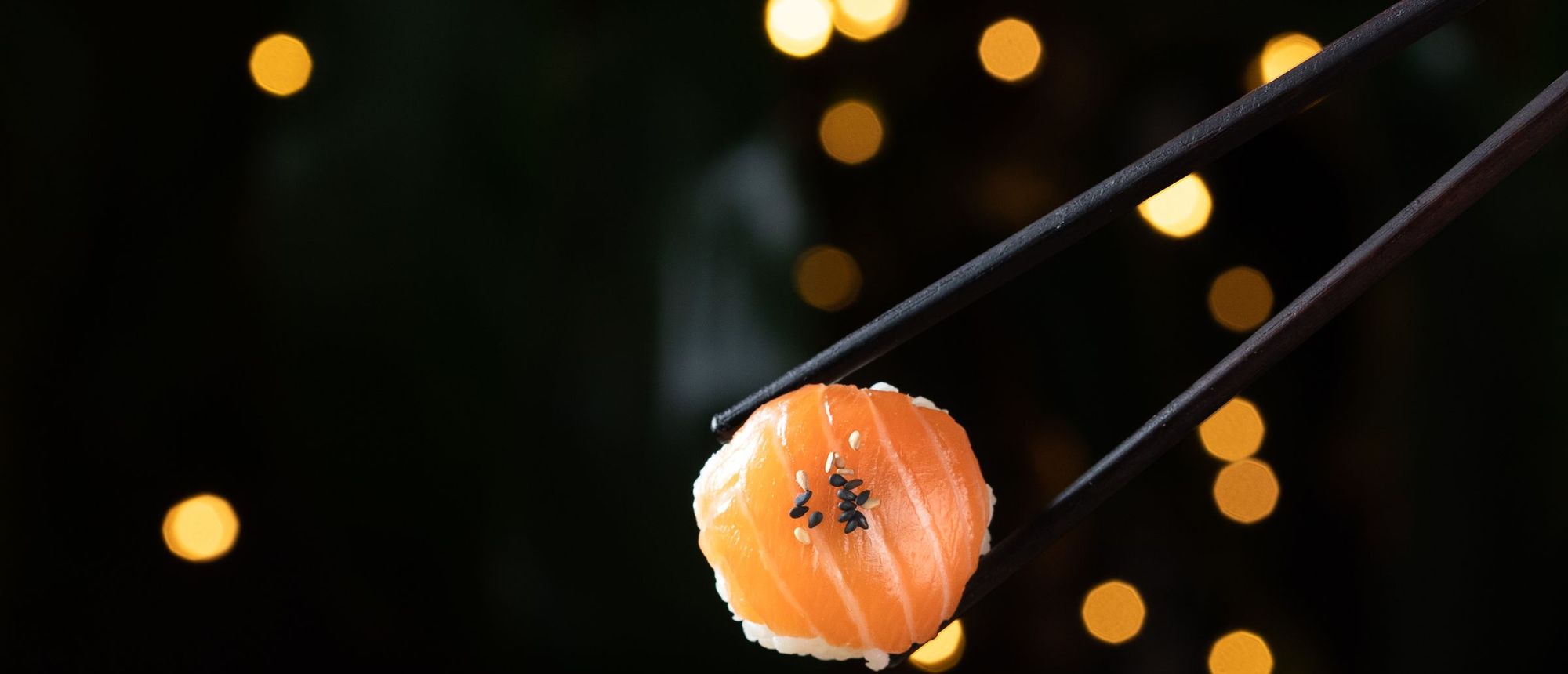 Chopsticks lifting a small ball of salmon temari sushi against warm evening lights