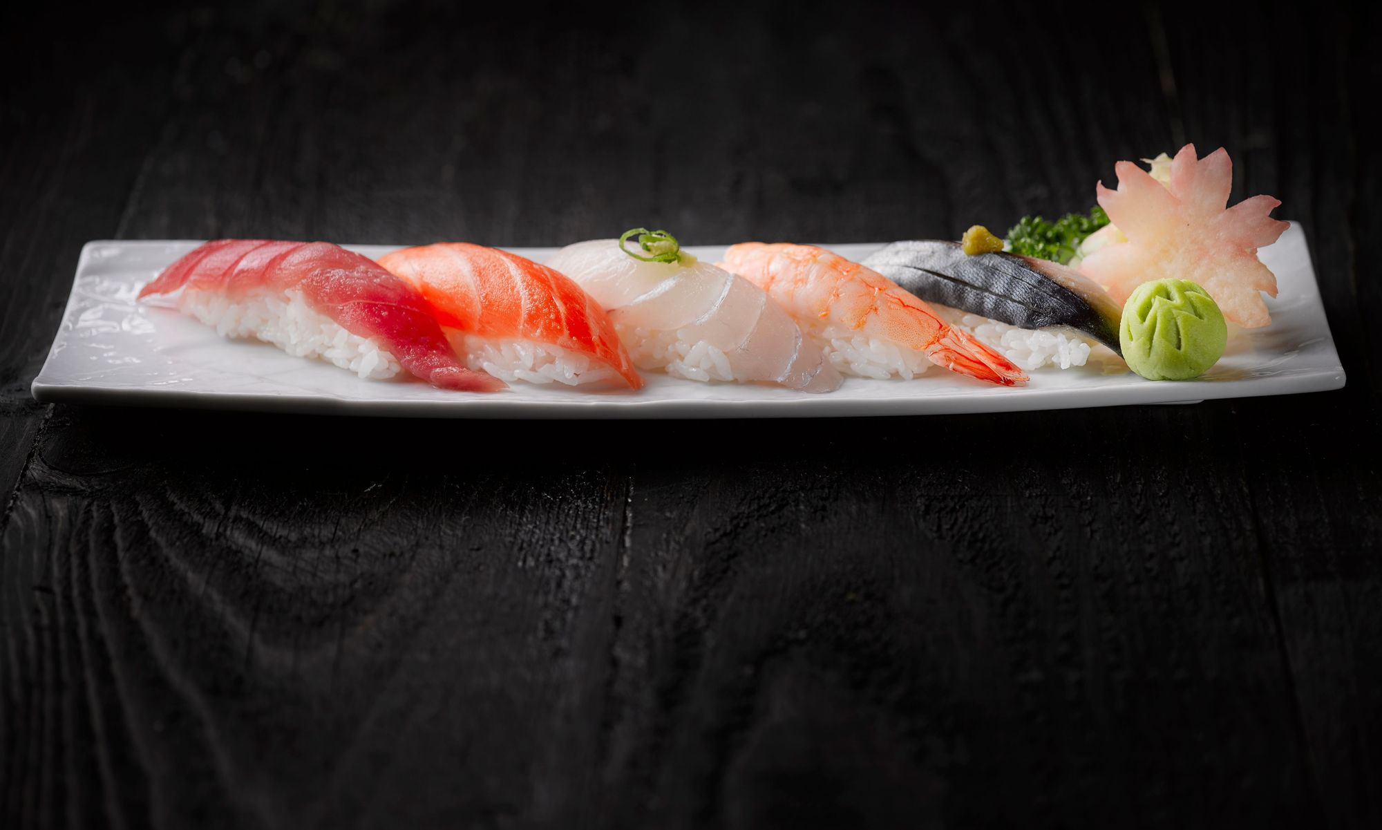 Five pieces of nigiri in a row on a long white plate, tuna, salmon, white fish, shrimp and mackerel, with wasabi and a pickled radish flower, on a black wooden counter
