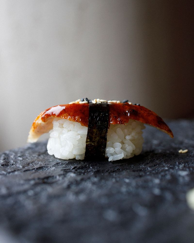 A piece of grilled eel nigiri with a band of nori, on a dark stone slab