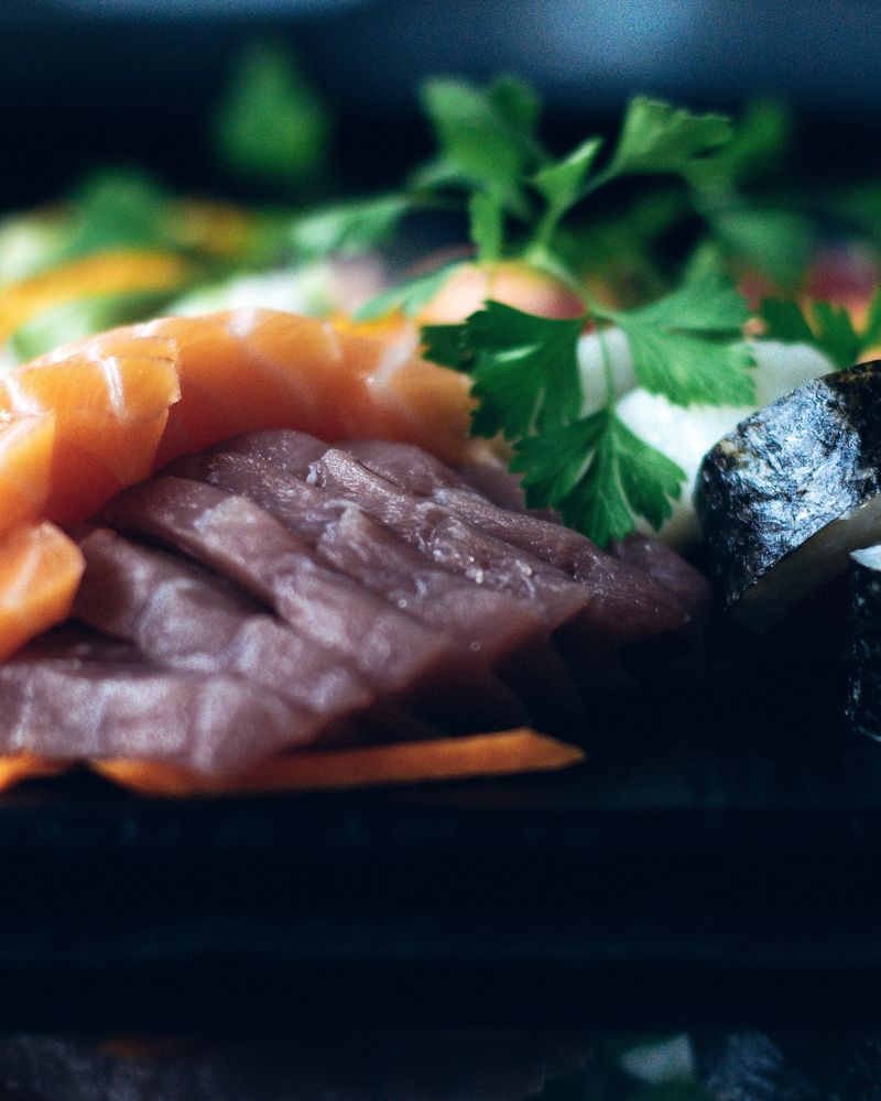 Folded slices of salmon and tuna sashimi beside a cut maki roll and chopsticks, on a black tray