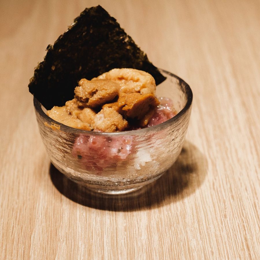 A small textured glass cup of chopped tuna heaped with sea urchin, a sheet of nori standing behind it, on a pale wood counter