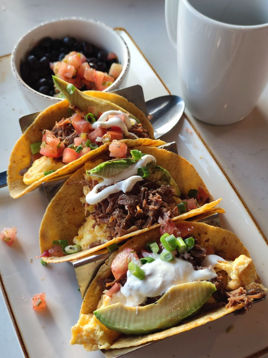 Three breakfast tacos filled with scrambled egg, shredded beef, avocado, pico de gallo and crema, with black beans and a mug of coffee