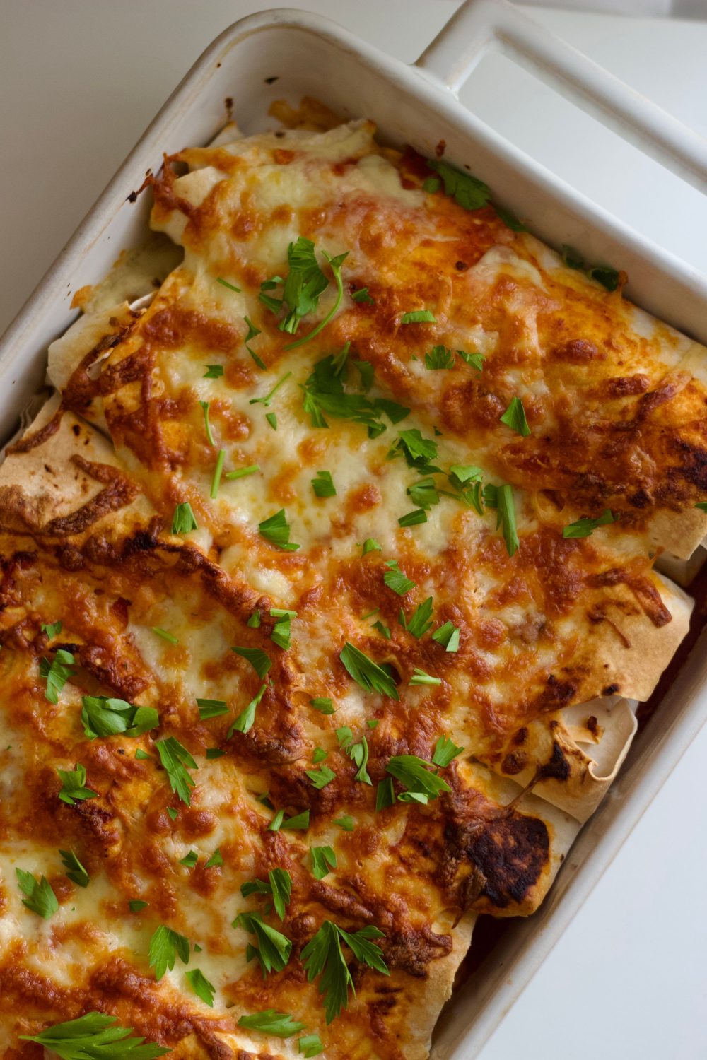A baking dish of enchiladas under browned cheese and red chile sauce, scattered with chopped herbs
