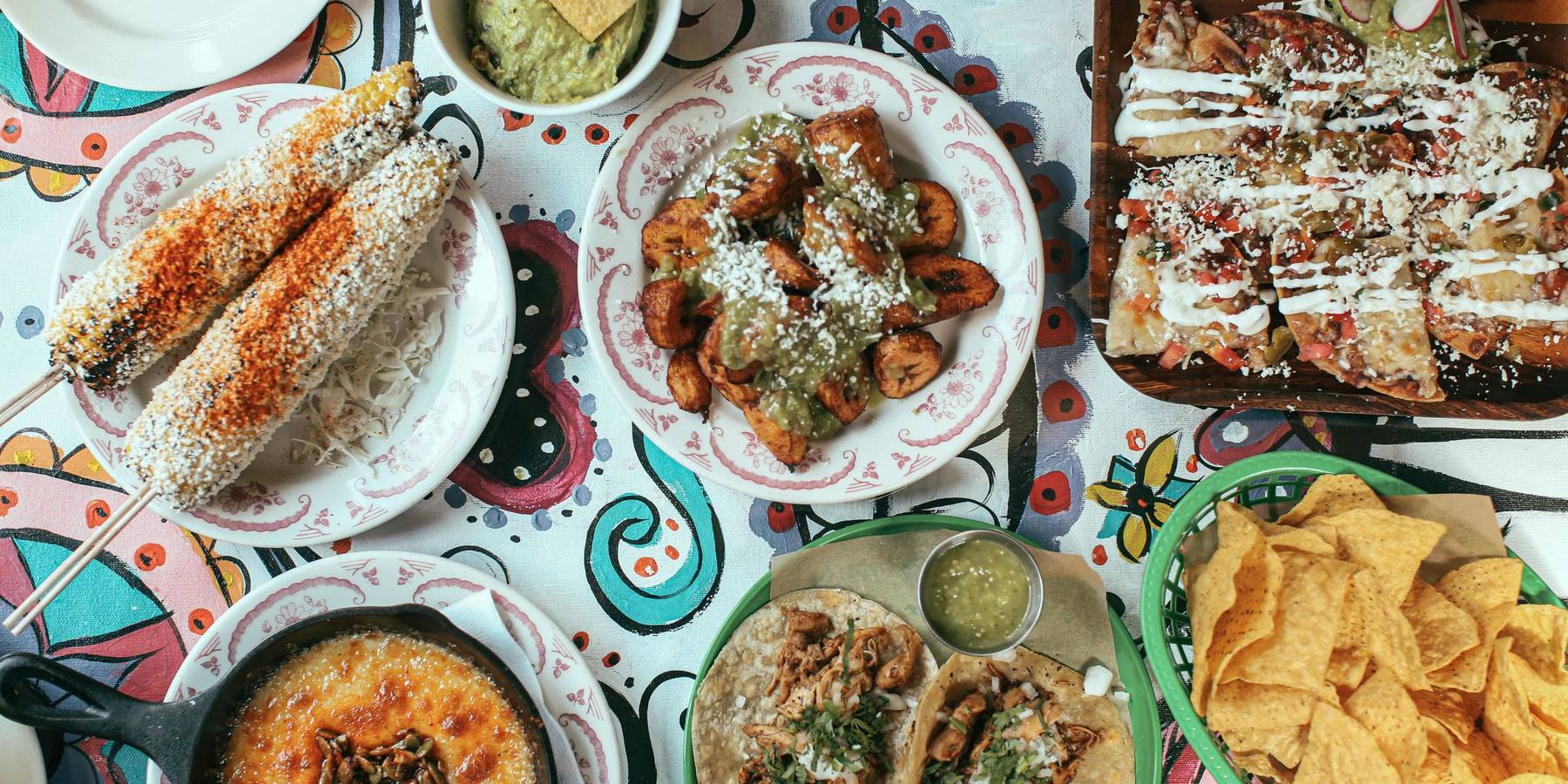 A table covered with elote, guacamole, fried plantains in salsa verde, tostadas, chicken tacos, queso fundido in a skillet and a basket of chips