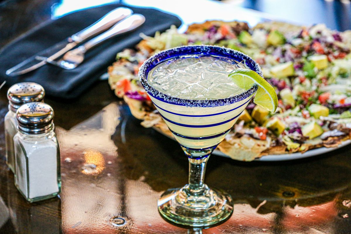 A salt rimmed margarita with a lime wedge in a blue striped glass, beside a platter of tostadas