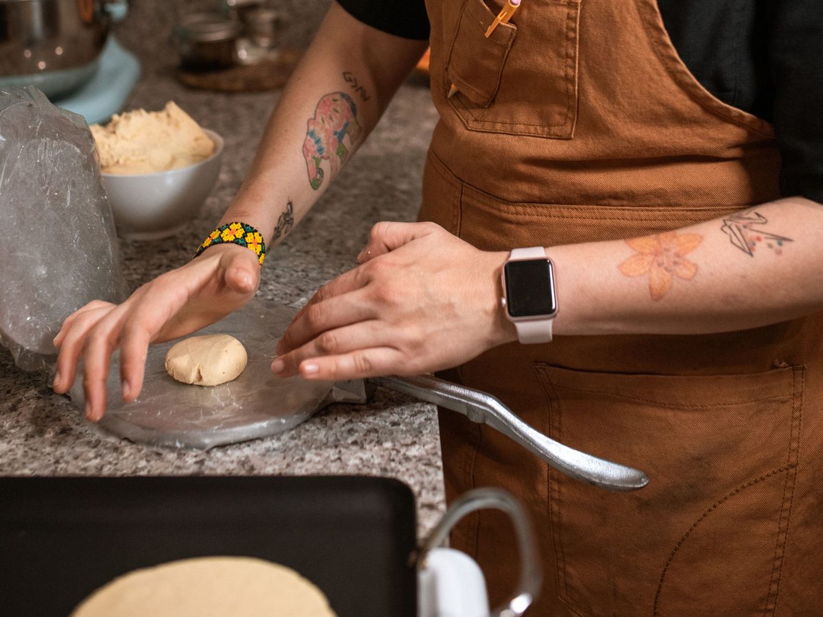 A cook's hands flattening a ball of masa on a tortilla press, with a bowl of dough and a hot griddle nearby