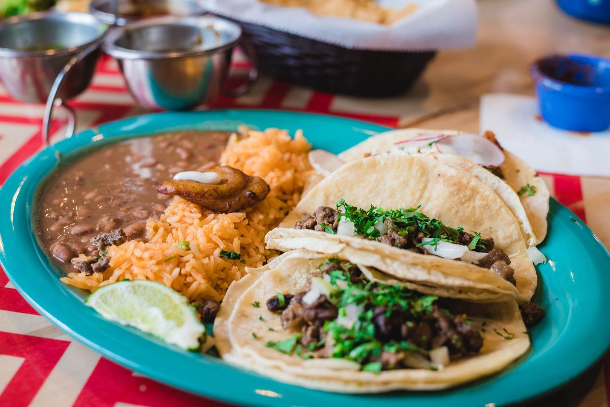 Beef tacos on corn tortillas with cilantro and onion, beside Mexican rice, pinto beans and a lime wedge on a turquoise plate